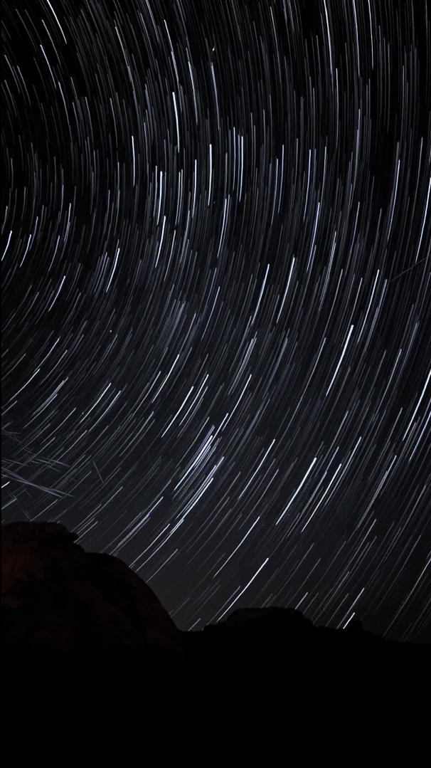 Long-exposure star trails curving above the dark silhouette of desert rocks.